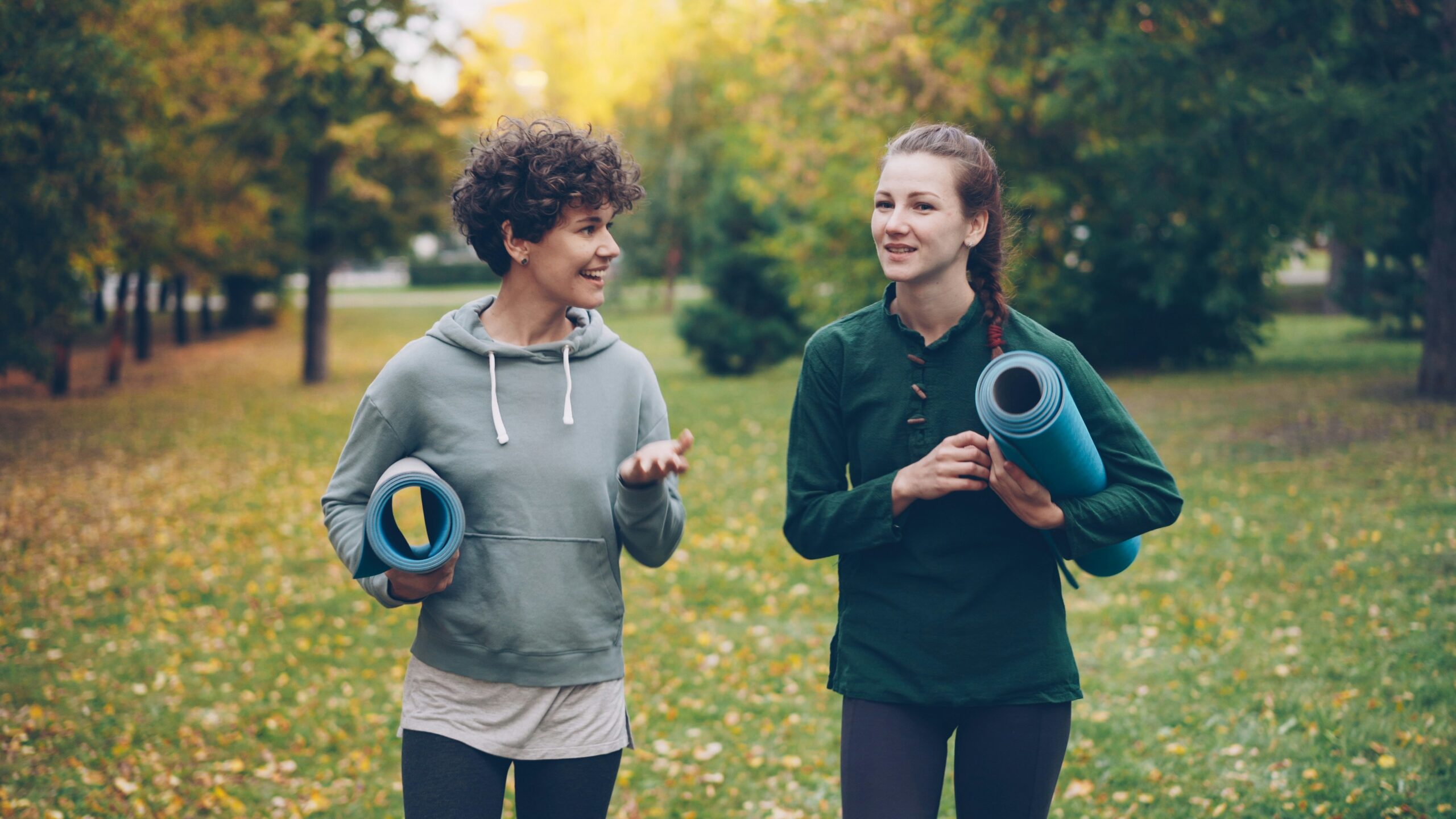 Two friends walking and talking in a park, holding rolled-up mats