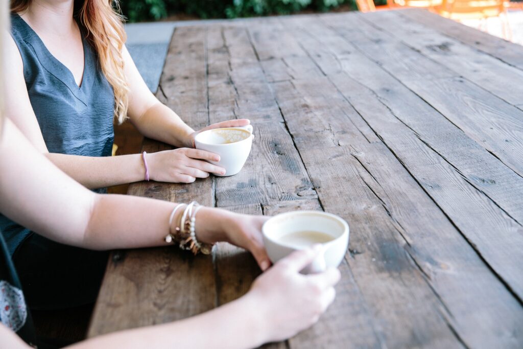 Two women sitting at an outdoor wooden table holding coffee cups and talking